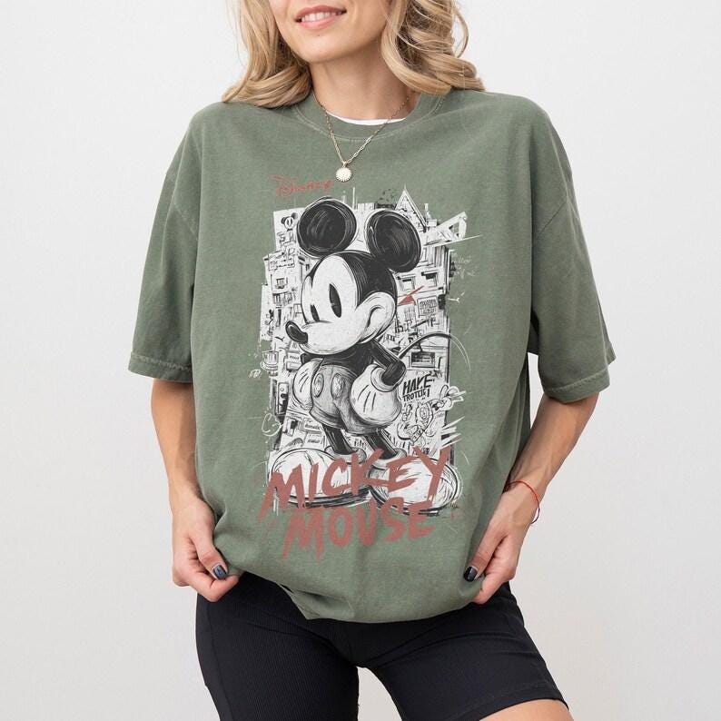 Puede incluir: Camiseta verde oliva con un gr&aacute;fico en blanco y negro de Mickey Mouse. El dise&ntilde;o incluye las palabras "Mickey Mouse" en rojo y un fondo de paisaje urbano. La camiseta tiene mangas cortas y un corte holgado.