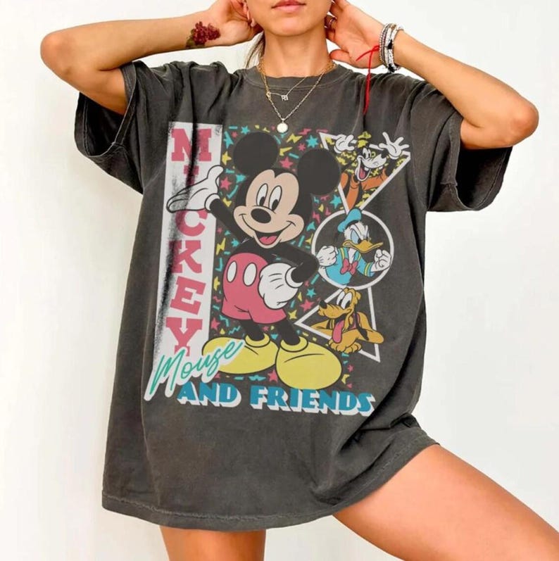 May include: Dark gray t-shirt featuring Mickey Mouse and friends. The graphic includes Mickey Mouse in a pose, with Donald Duck and Goofy. The text "Mickey Mouse AND FRIENDS" is displayed on the front.