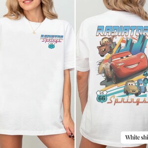 May include: A white t-shirt featuring a small Radiator Springs US 66 logo on the front chest. The back displays a large graphic of Lightning McQueen, Mater, and other characters with the text Radiator Springs and a US 66 road sign emblem.