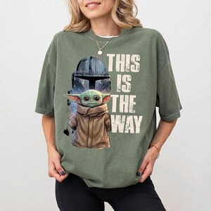 May include: Green t-shirt with a graphic of a character from a popular science fiction series. The design includes a helmet, a small green creature, and the text "THIS IS THE WAY" in white.