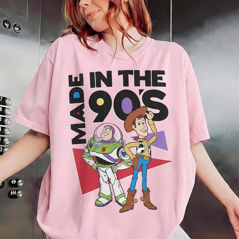 May include: Pink t-shirt with the text "MADE IN THE 90'S" and cartoon images of Buzz Lightyear and Woody. The shirt has a crew neck and short sleeves. The graphic features a retro design with a red and purple triangle.