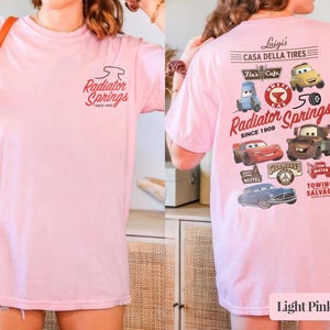 May include: A light pink t-shirt featuring a small Radiator Springs logo on the front. The back displays a large graphic with various cartoon car characters, the text Luigi's Casa Della Tires, Radiator Springs Since 1909, and Tow Mater.