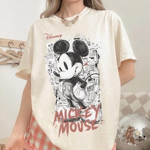 Puede incluir: Camiseta color crema con un gr&aacute;fico en blanco y negro de Mickey Mouse, la palabra "Disney" arriba y "Mickey Mouse" en rojo abajo. El dise&ntilde;o incluye un fondo de paisaje urbano.