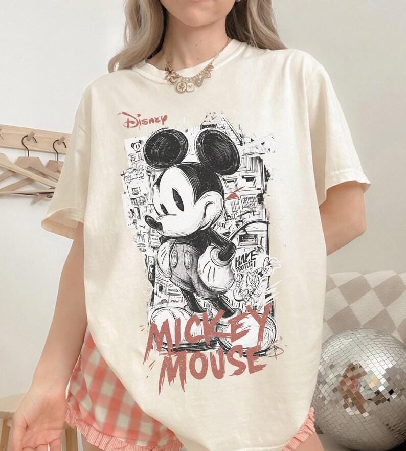 Puede incluir: Camiseta color crema con un gr&aacute;fico en blanco y negro de Mickey Mouse, la palabra "Disney" arriba y "Mickey Mouse" en rojo abajo. El dise&ntilde;o incluye un fondo de paisaje urbano.