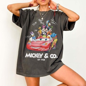 May include: Dark gray t-shirt featuring a graphic of Disney characters including Mickey Mouse, Minnie Mouse, Donald Duck, and Lightning McQueen. The shirt has the text "Mickey & Co. EST 1928" and character signatures.