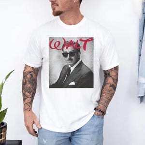 May include: White t-shirt featuring a black and white portrait of a man wearing sunglasses and a suit. The word "WALT" is printed in red cursive above the portrait. The shirt is made of a soft material.