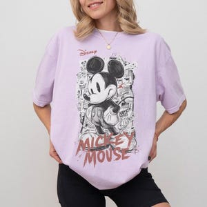 Puede incluir: Camiseta lila claro con un gr&aacute;fico en blanco y negro de Mickey Mouse. El dise&ntilde;o incluye el texto "Disney" y "Mickey Mouse" en una fuente desgastada. La camiseta tiene mangas cortas y un corte holgado.