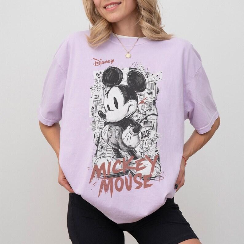 Puede incluir: Camiseta lila claro con un gr&aacute;fico en blanco y negro de Mickey Mouse. El dise&ntilde;o incluye el texto "Disney" y "Mickey Mouse" en una fuente desgastada. La camiseta tiene mangas cortas y un corte holgado.