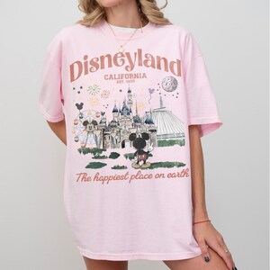 Puede incluir: Una camiseta rosa claro con un gr&aacute;fico de Disneyland. El dise&ntilde;o incluye el logotipo de Disneyland, un castillo, Mickey Mouse y el texto "The happiest place on earth."