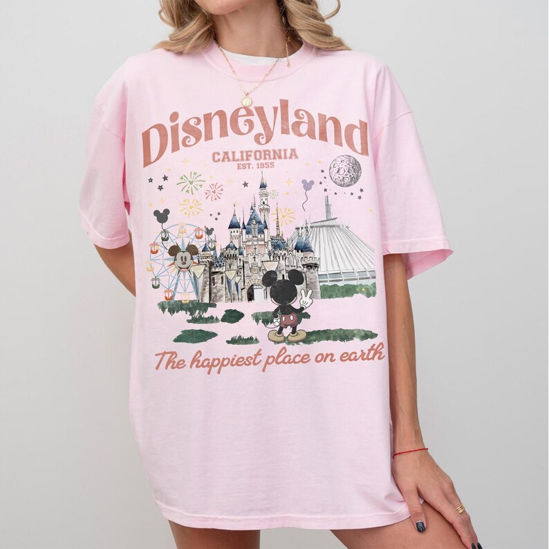 Puede incluir: Una camiseta rosa claro con un gr&aacute;fico de Disneyland. El dise&ntilde;o incluye el logotipo de Disneyland, un castillo, Mickey Mouse y el texto "The happiest place on earth."