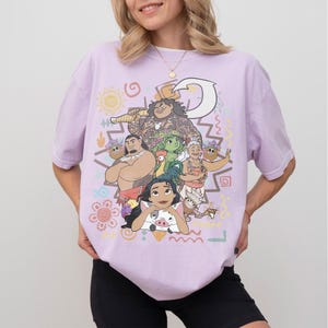 May include: A light purple short-sleeve t-shirt featuring a colorful graphic print of various animated characters, including a large figure holding a hook, a pig, a chicken, and several other figures, surrounded by small decorative shapes and symbols.