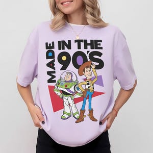 May include: A light purple t-shirt featuring a graphic of Buzz Lightyear and Woody from Toy Story, with the text "MADE IN THE 90'S". The design includes red, purple, and white geometric shapes. The shirt has short sleeves.