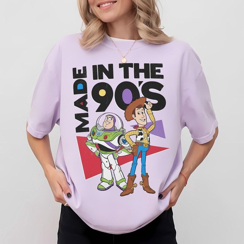 May include: A light purple t-shirt featuring a graphic of Buzz Lightyear and Woody from Toy Story, with the text "MADE IN THE 90'S". The design includes red, purple, and white geometric shapes. The shirt has short sleeves.