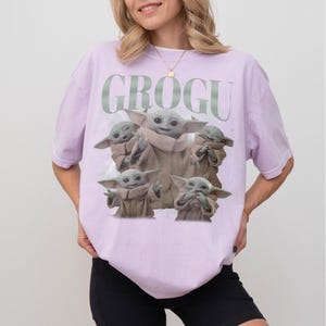 May include: A light purple t-shirt featuring a graphic of Grogu from The Mandalorian. The design includes multiple images of Grogu in various poses, with the word "GROGU" printed above. The shirt is a relaxed fit.
