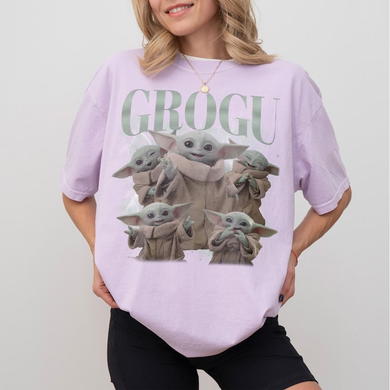May include: A light purple t-shirt featuring a graphic of Grogu from The Mandalorian. The design includes multiple images of Grogu in various poses, with the word "GROGU" printed above. The shirt is a relaxed fit.