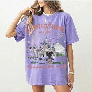 Puede incluir: Una camiseta lila con un gr&aacute;fico de Disneyland. El dise&ntilde;o incluye el logotipo de Disneyland, la ubicaci&oacute;n en California y la frase "The happiest place on earth". El gr&aacute;fico tambi&eacute;n muestra un castillo, Mickey Mouse y otros elementos del parque.