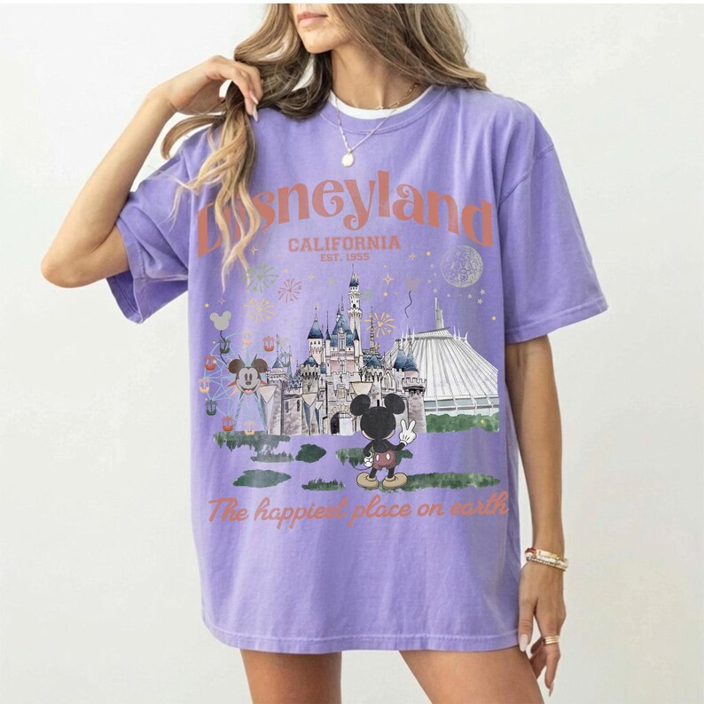 Puede incluir: Una camiseta lila con un gr&aacute;fico de Disneyland. El dise&ntilde;o incluye el logotipo de Disneyland, la ubicaci&oacute;n en California y la frase "The happiest place on earth". El gr&aacute;fico tambi&eacute;n muestra un castillo, Mickey Mouse y otros elementos del parque.