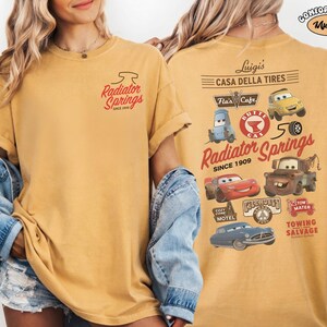 May include: A mustard yellow t-shirt featuring a small Radiator Springs logo on the front left chest. The back displays a large graphic with various Cars movie characters, including Lightning McQueen, Mater, and Luigi, alongside text like Casa Della Tires.