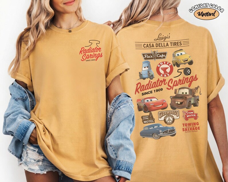 May include: A mustard yellow t-shirt featuring a small Radiator Springs logo on the front left chest. The back displays a large graphic with various Cars movie characters, including Lightning McQueen, Mater, and Luigi, alongside text like Casa Della Tires.