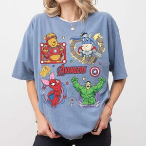 Puede incluir: Camiseta azul claro con personajes de dibujos animados de los Vengadores. La camiseta presenta a Iron Man, Thor, Spider-Man y Hulk, con la palabra "Avengers" en el centro.