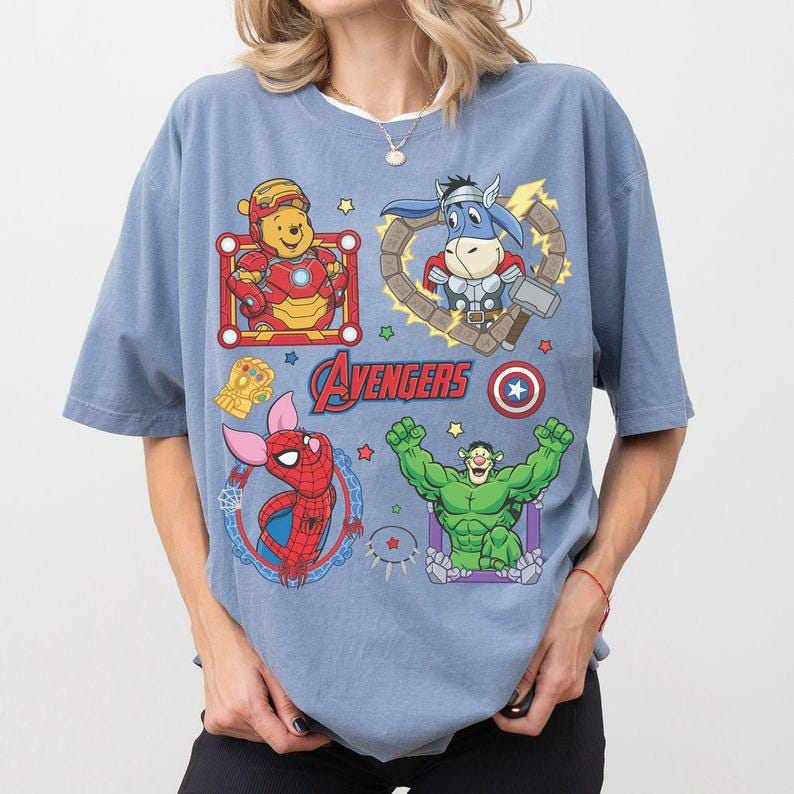 Puede incluir: Camiseta azul claro con personajes de dibujos animados de los Vengadores. La camiseta presenta a Iron Man, Thor, Spider-Man y Hulk, con la palabra "Avengers" en el centro.