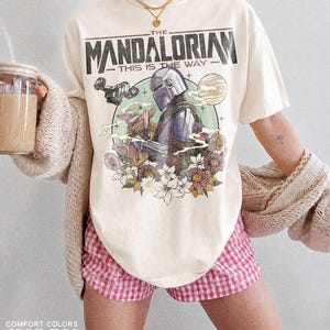 May include: Cream-coloured t-shirt with a graphic of The Mandalorian and Grogu, encircled by flowers. The text "THE MANDALORIAN - THIS IS THE WAY" is printed across the top. Part of the Comfort Colors collection.