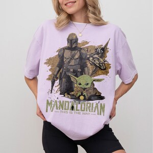 May include: Lilac-colored t-shirt featuring a graphic of The Mandalorian and Grogu. The design includes a spaceship and the text "MANDALORIAN - THIS IS THE WAY" in a stylized font. The shirt has short sleeves and a relaxed fit.