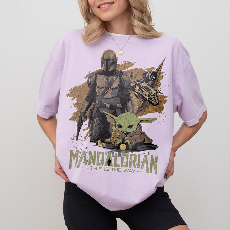 May include: Lilac-colored t-shirt featuring a graphic of The Mandalorian and Grogu. The design includes a spaceship and the text "MANDALORIAN - THIS IS THE WAY" in a stylized font. The shirt has short sleeves and a relaxed fit.