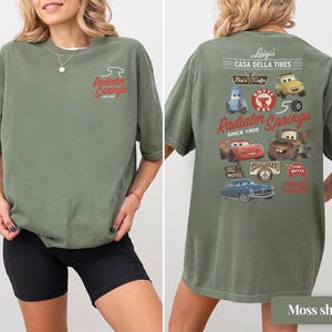 May include: An olive green t-shirt featuring a small Radiator Springs logo on the front chest. The back displays a large graphic with various Cars characters, including Lightning McQueen and Mater, alongside text reading Luigi's Casa Della Tires.
