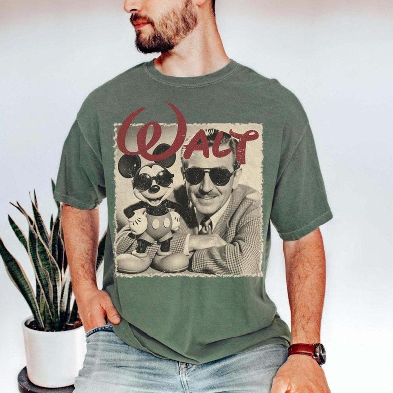 May include: Green t-shirt featuring a vintage-style graphic of Walt Disney and Mickey Mouse wearing sunglasses. The word "Walt" is printed in a red, stylized font above the image. The shirt has short sleeves and a crew neck.