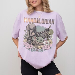 May include: A light purple t-shirt featuring the text "MANDALORIAN" above a graphic of the Mandalorian, Grogu, and a spaceship, surrounded by flowers. The shirt is a relaxed fit.