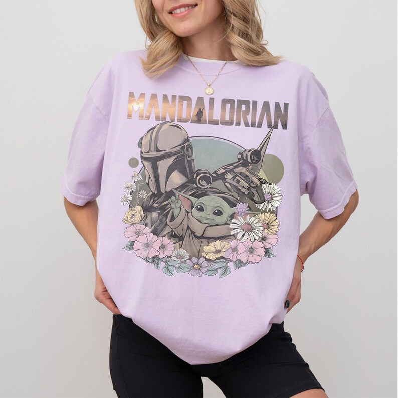 May include: A light purple t-shirt featuring the text "MANDALORIAN" above a graphic of the Mandalorian, Grogu, and a spaceship, surrounded by flowers. The shirt is a relaxed fit.