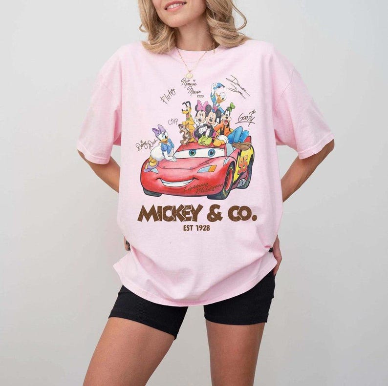 May include: Pink t-shirt featuring a graphic of Mickey & Co. characters in a red car. The design includes signatures and the text "Mickey & Co. EST 1928". The shirt has short sleeves and a relaxed fit.