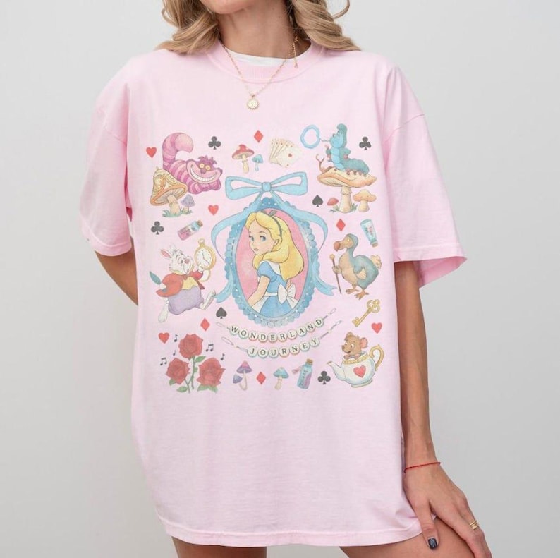 May include: Pink t-shirt featuring an Alice in Wonderland design. The graphic includes Alice, the Cheshire Cat, and other characters. The words "Wonderland Journey" are written below the central image. The shirt has short sleeves and a crew neck.