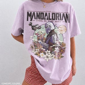 May include: A lilac t-shirt featuring a graphic from "The Mandalorian." The design includes the Mandalorian, Grogu, a blaster, and a sun, framed by flowers. The text "THE MANDALORIAN" and "THIS IS THE WAY" are printed above the image.