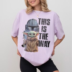 May include: A lilac t-shirt with a graphic of Grogu, the Mandalorian helmet, and the text "THIS IS THE WAY" in black. The Grogu illustration is in a tan coat. The shirt has a loose fit.