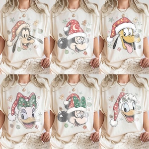 Peut inclure: Une collection de six t-shirts couleur crème présentant divers personnages Disney portant des bonnets de Père Noël rouges et blancs. Les motifs incluent Dingo, Mickey, Pluto, Daisy et Donald, entourés de bonshommes en pain d'épice et flocons.
