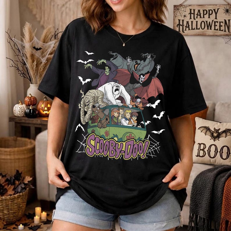 May include: A black t-shirt featuring a graphic print of the Scooby-Doo characters inside the Mystery Machine van, surrounded by various classic monsters including a mummy, a vampire, and a yeti, with white bats and spiderwebs in the background.