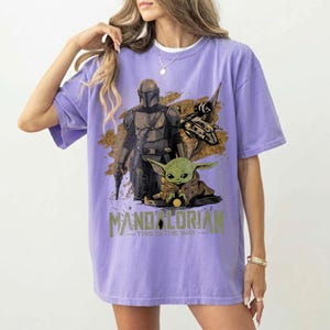 May include: A light purple t-shirt featuring a graphic of The Mandalorian, Grogu, and a spaceship. The text "MANDALORIAN" and "THIS IS THE WAY" are printed below the characters. The shirt is a comfortable, oversized fit.