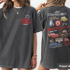 May include: A charcoal gray t-shirt featuring a small Radiator Springs logo on the front chest. The back displays a large graphic with various Cars movie characters, including Lightning McQueen and Mater, alongside text like Luigi's Casa Della Tires.