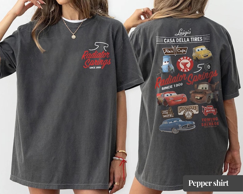 May include: A charcoal gray t-shirt featuring a small Radiator Springs logo on the front chest. The back displays a large graphic with various Cars movie characters, including Lightning McQueen and Mater, alongside text like Luigi's Casa Della Tires.