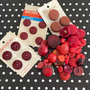 May include: A selection of red buttons in diverse forms, such as round, square, and floral. Some buttons remain on their original packaging, with the brand name "Beutron" displayed. The backdrop is a black surface adorned with white polka dots.