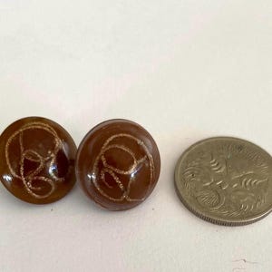 May include: Two brown, round earrings with a gold swirl design. A silver coin is next to the earrings for size comparison. The earrings have a glossy finish and are set against a white background.
