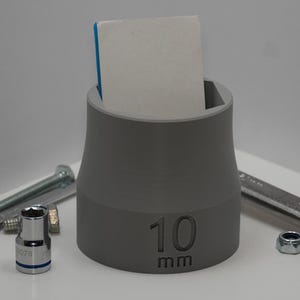 May include: A gray 10 mm socket holder with a white and blue object inside, surrounded by various tools including a wrench, bolts, and nuts. The holder is designed to organize and store sockets, and the number "10 mm" is printed on the side.