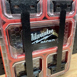 Puede incluir: Primer plano de un organizador de herramientas de plástico rojo y transparente con soportes metálicos negros. El organizador presenta un patrón de panal y el logotipo de Milwaukee PACKOUT es visible.