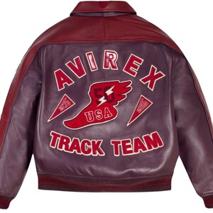 Pode incluir: Casaco de couro bord&ocirc; com gola e mangas marrons. As costas apresentam o texto "AVIREX TRACK TEAM" em letras brancas com detalhes em vermelho. Tamb&eacute;m est&aacute; presente um gr&aacute;fico vermelho e branco de um p&eacute; alado e um raio.