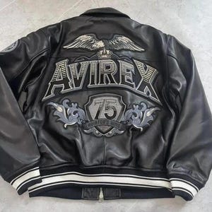 May include: Black leather jacket with white and black striped trim. The back features a large embroidered design with the word "AVIREX", an eagle, and the text "75 Aviator's King". A zipper runs down the front.