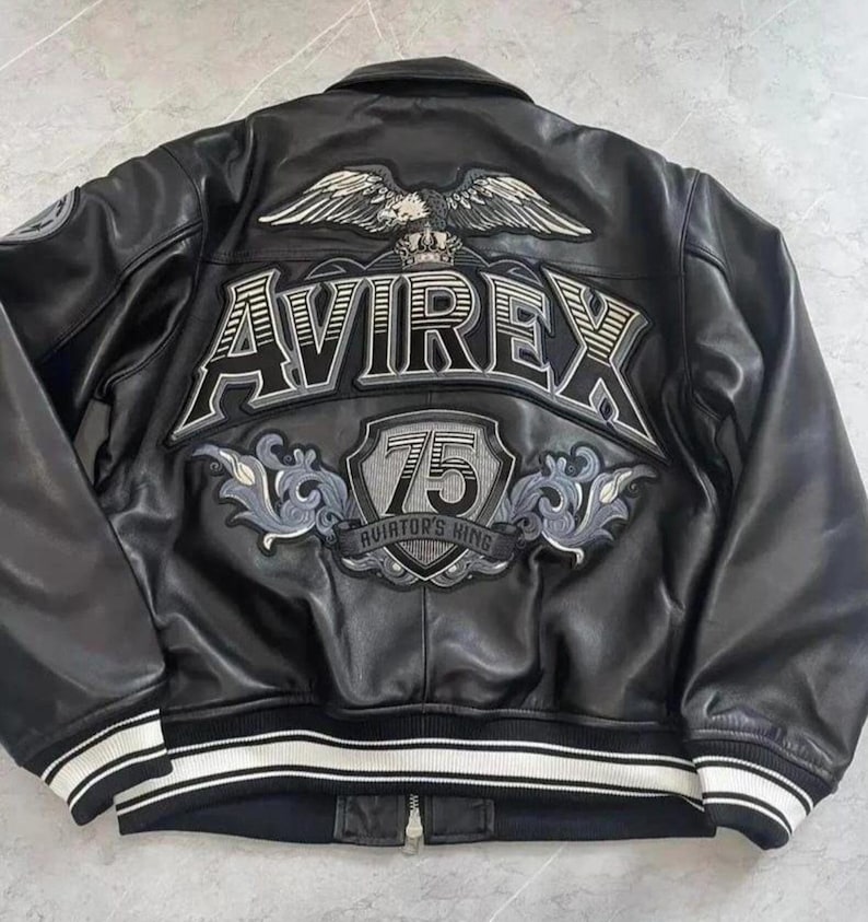 May include: Black leather jacket with white and black striped trim. The back features a large embroidered design with the word "AVIREX", an eagle, and the text "75 Aviator's King". A zipper runs down the front.