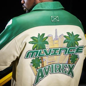 May include: A green and cream-colored leather jacket with a city and palm tree graphic on the back. The jacket features the brand name "MLVINCE AVIREX" in green lettering. The jacket has a green collar and shoulder accents.