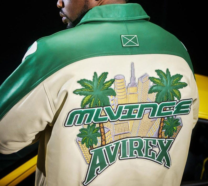 May include: A green and cream-colored leather jacket with a city and palm tree graphic on the back. The jacket features the brand name "MLVINCE AVIREX" in green lettering. The jacket has a green collar and shoulder accents.